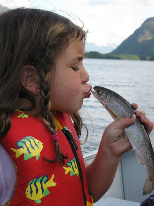 A child poses with a small fish, pretending to kiss the fish. Calm waters, sunny skies and a tree covered mountain are visible behind them.